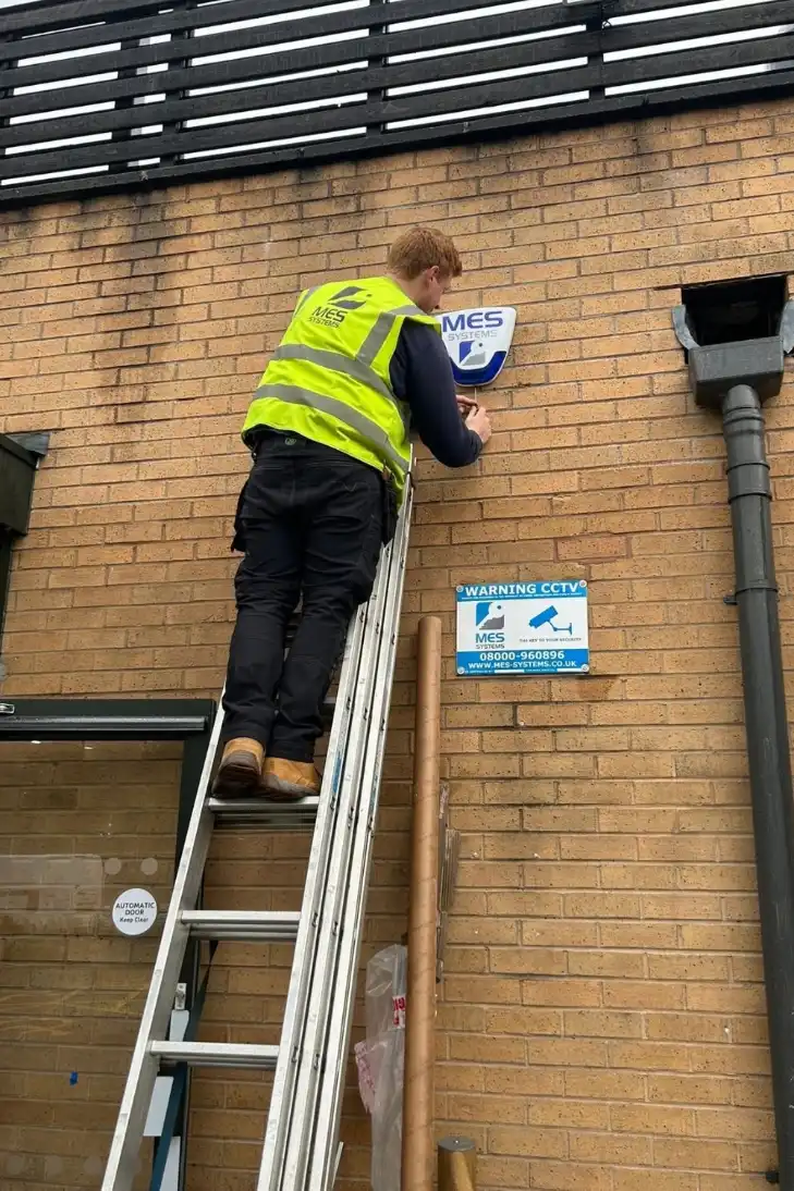 MES Systems engineer fitting an alarm bell box to a brick wall from a ladder